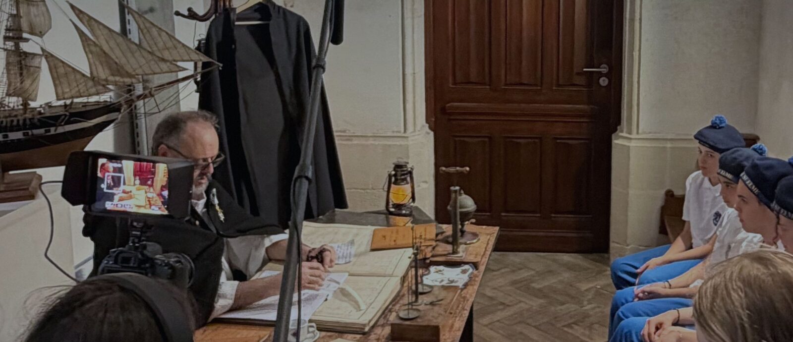 Sur le plateau en costume d'époque : un comédien écrit à une table éclairée par une lanterne, des élèves en bérets de marin assis à droite, la caméra et la perche au premier plan
